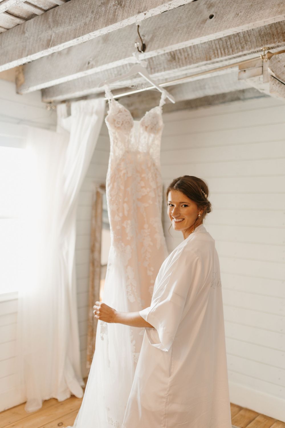 Smiling bride in white robe standing beside hanging lace wedding gown in bright rustic dressing room.