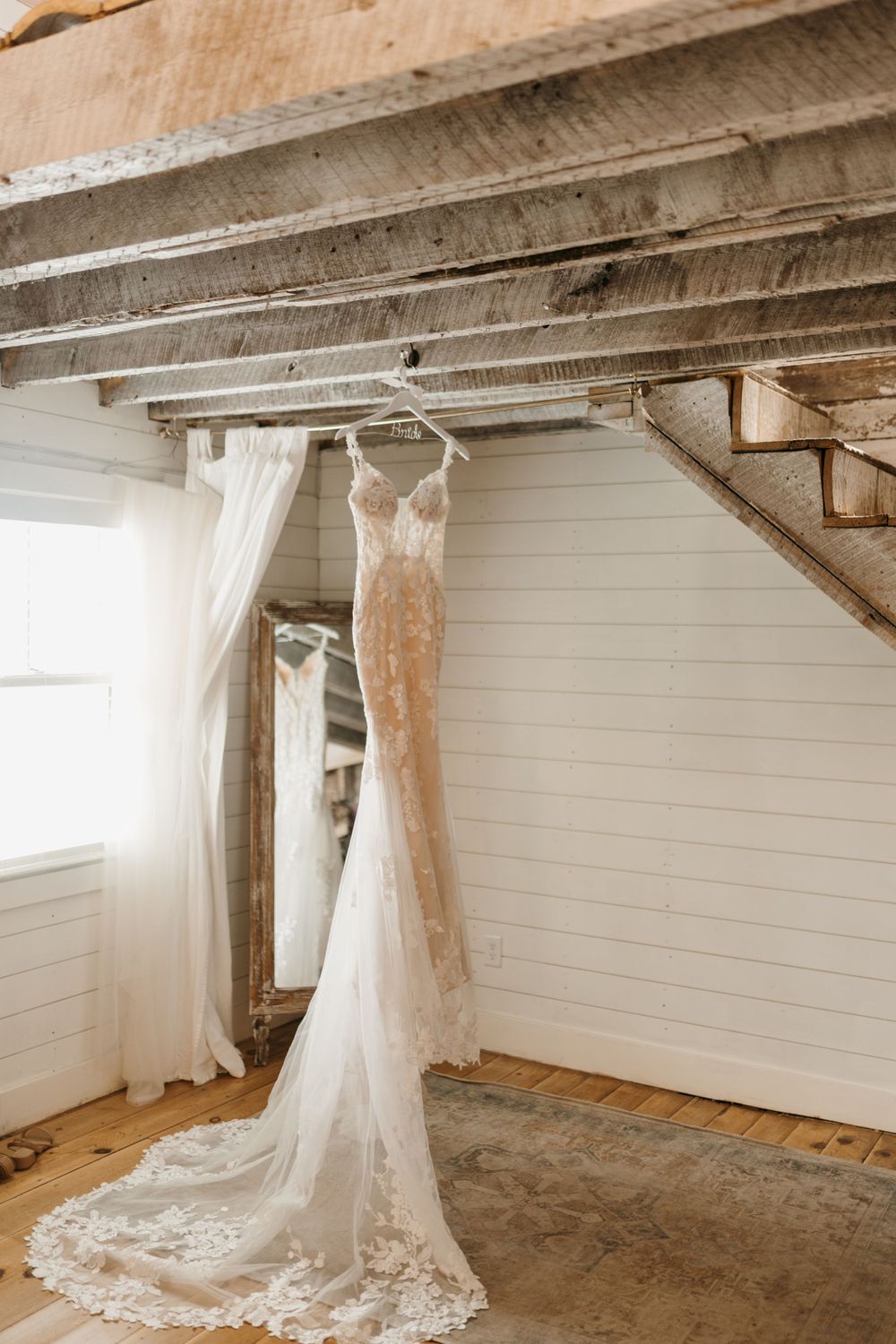 Elegant lace wedding gown hangs from rustic wooden beams in a bright white shiplap bridal suite.