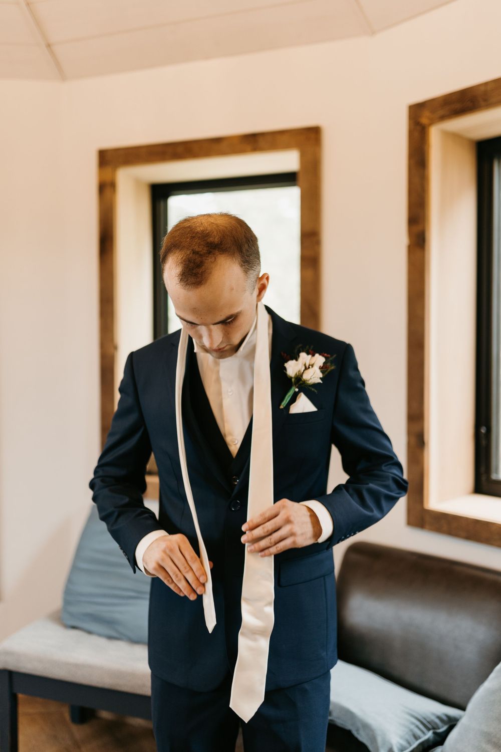 Groom in navy suit adjusting cream tie with boutonniere before wedding ceremony.