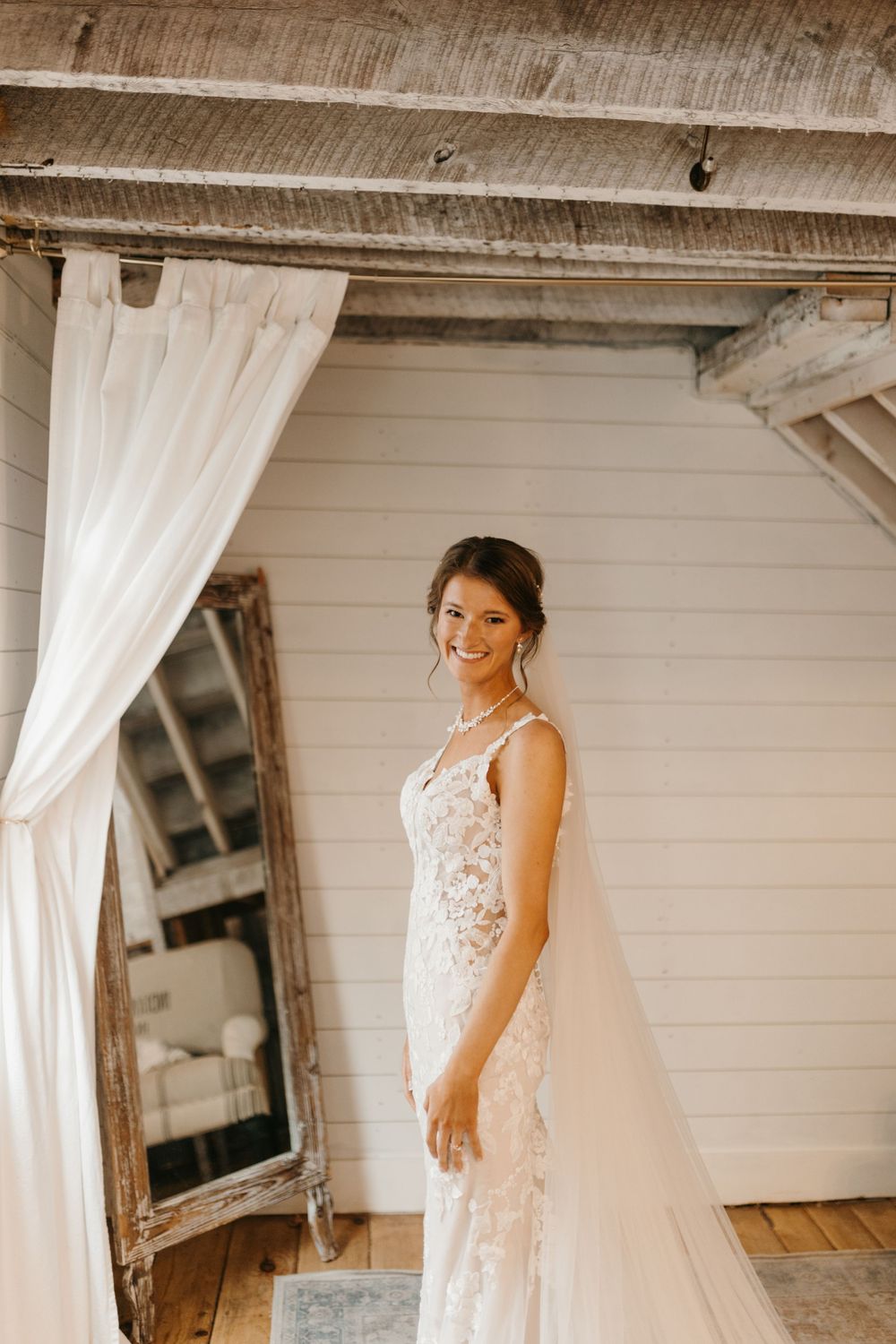 Radiant bride in fitted lace wedding gown with train posing beside white curtains in rustic venue.