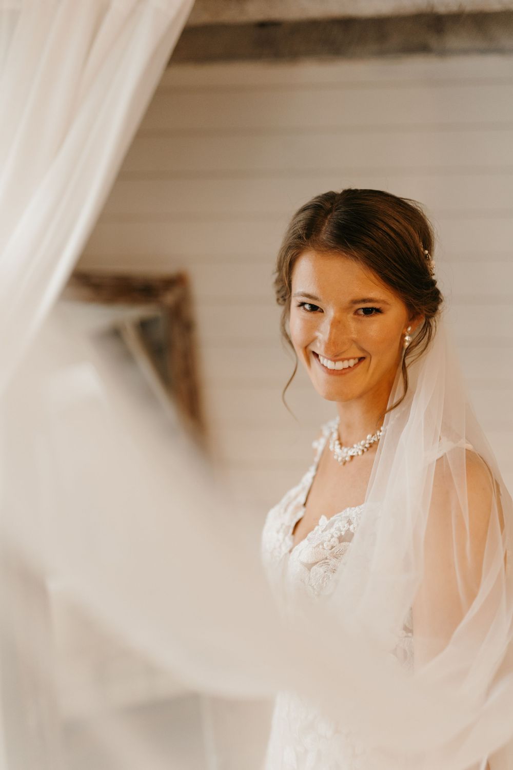 Glowing bride in lace gown and veil smiling softly through sheer white curtains in natural light.