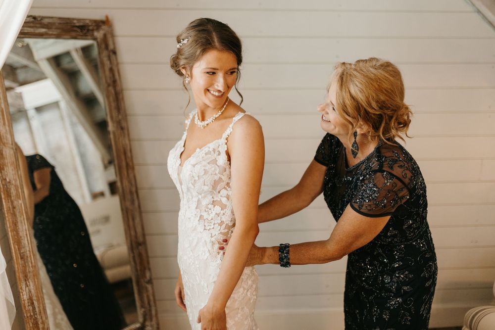 Bride smiling as woman in navy sequin dress adjusts her lace wedding gown beside ornate mirror.