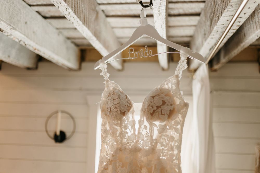 Lace wedding dress hangs on a personalized Bride hanger beneath rustic wooden beams in a charming venue.