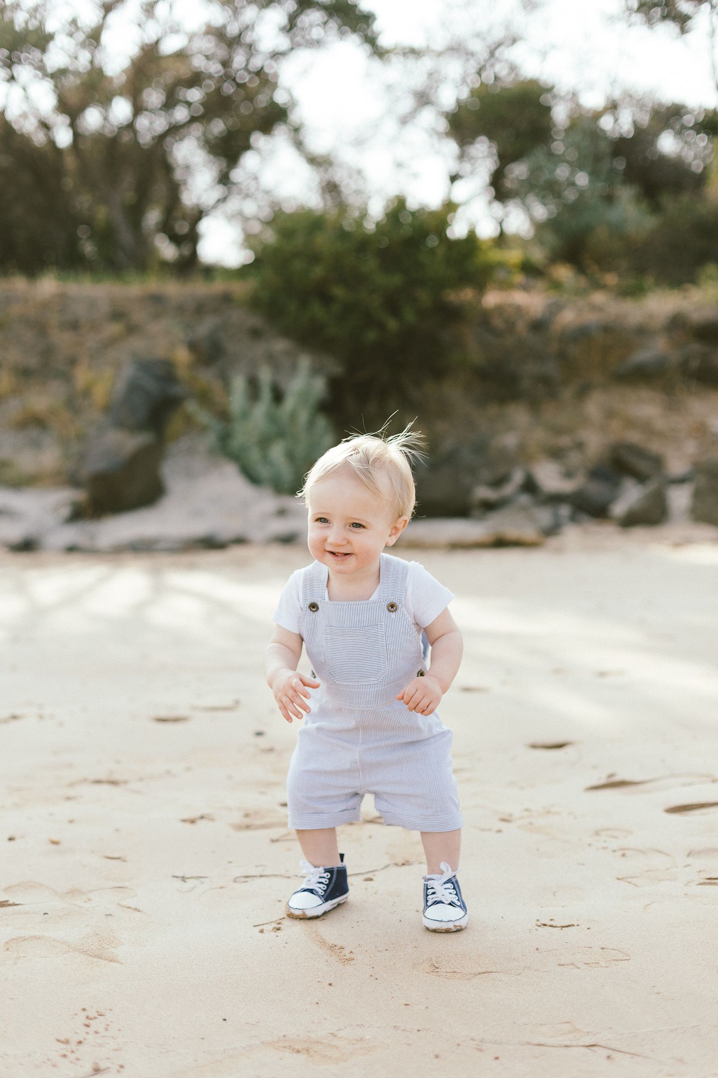 A baby in white clothing crawls and plays on a sandy beach with greenery in the background.