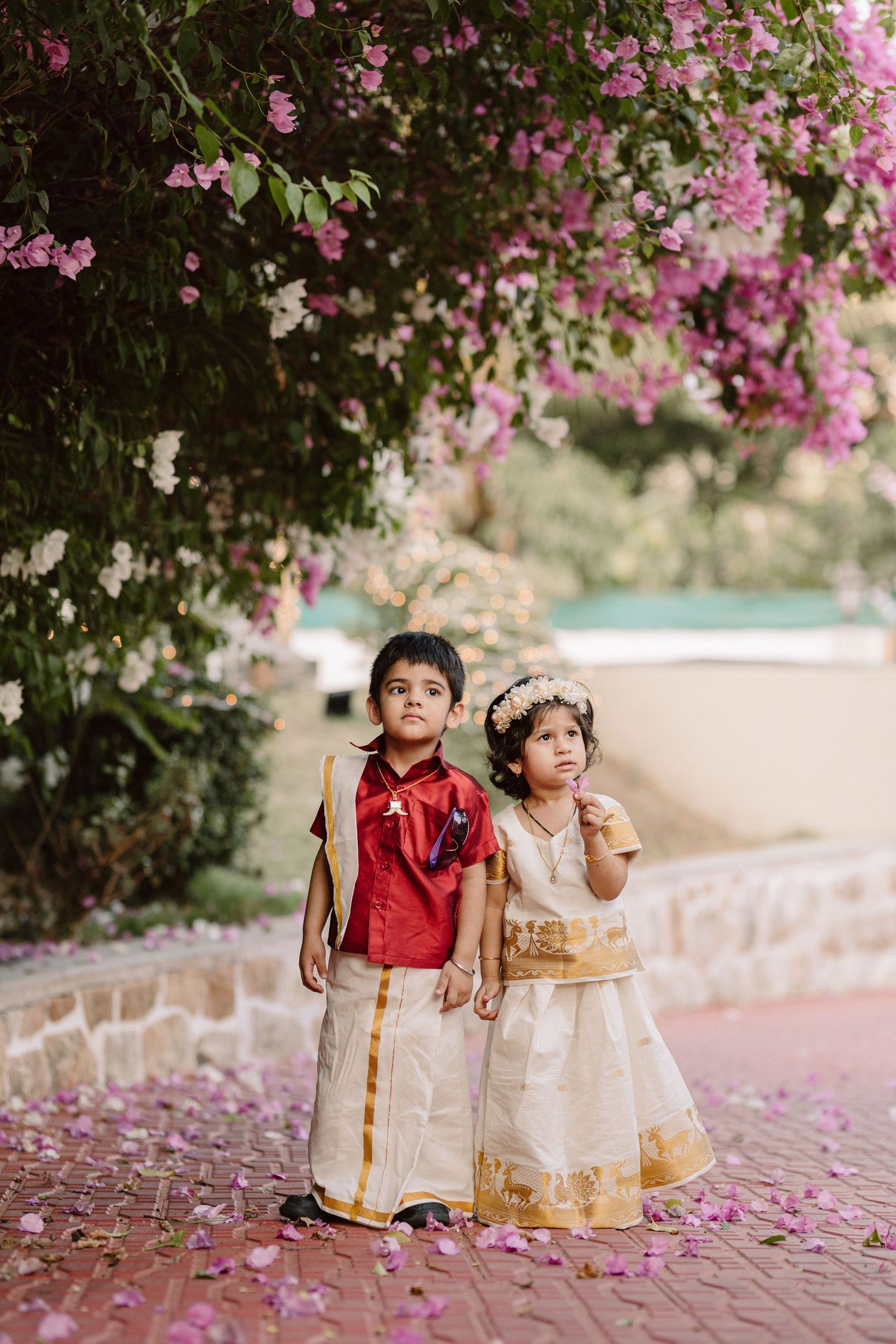 Two small children in traditional Indian attire sitting together under blooming pink bougainvillea.