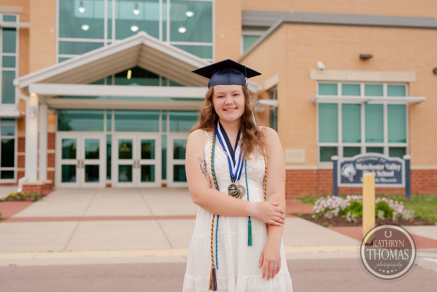 Graduate stands confidently in white dress and graduation regalia in front of school entrance.