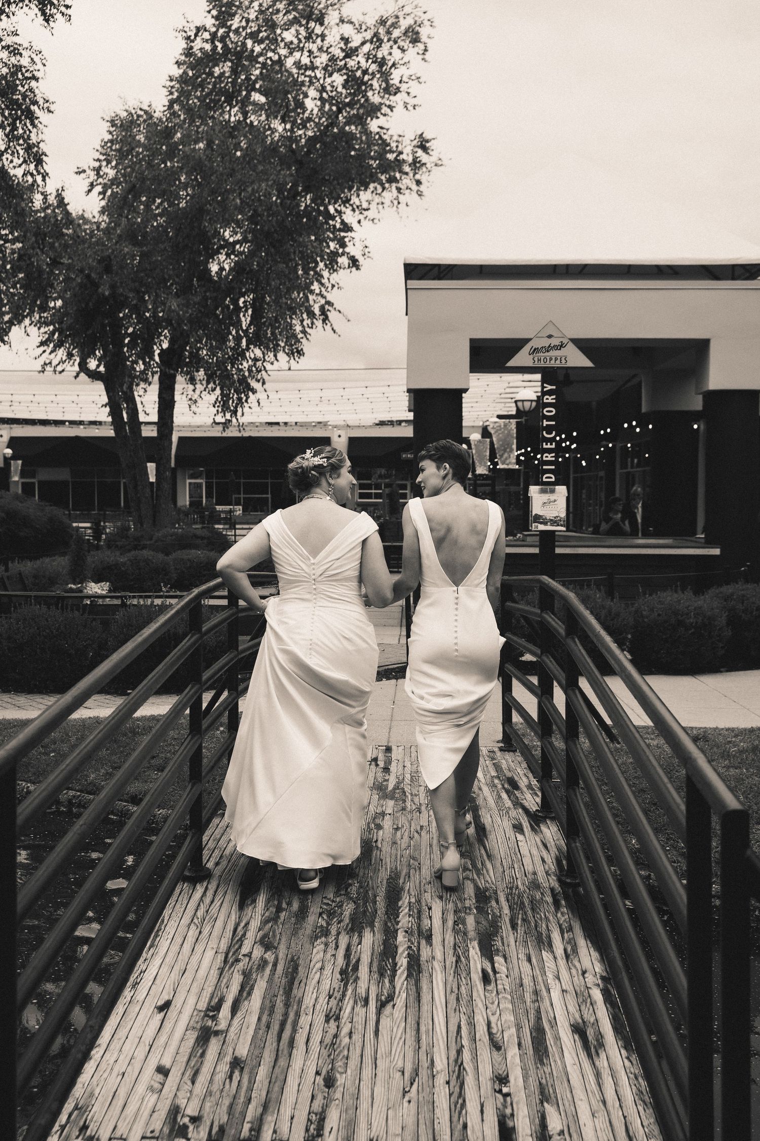 Black and white photo of two brides walking across the bridge showcasing the button details of their dresses.