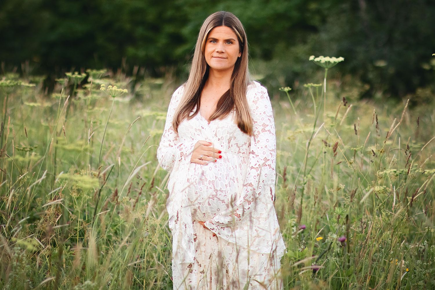 Maternity portrait in white lace dress amid wildflower field at golden hour.