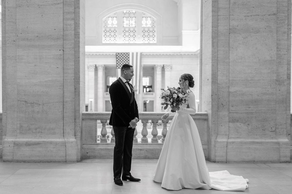 Black and white portrait of newlyweds standing by ornate window in historic building.