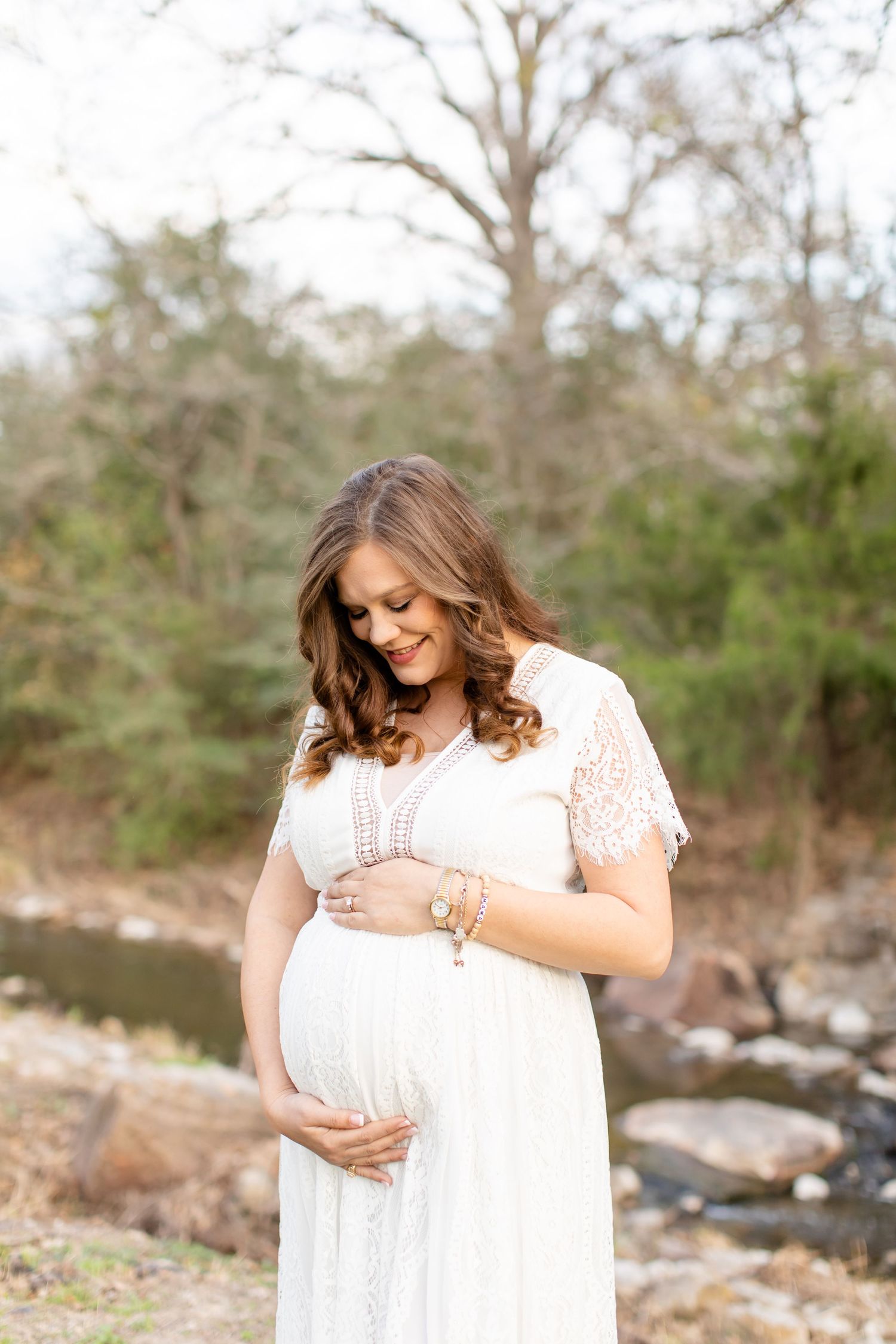 A woman in white dress looks down at her baby bump while standing by a stream.