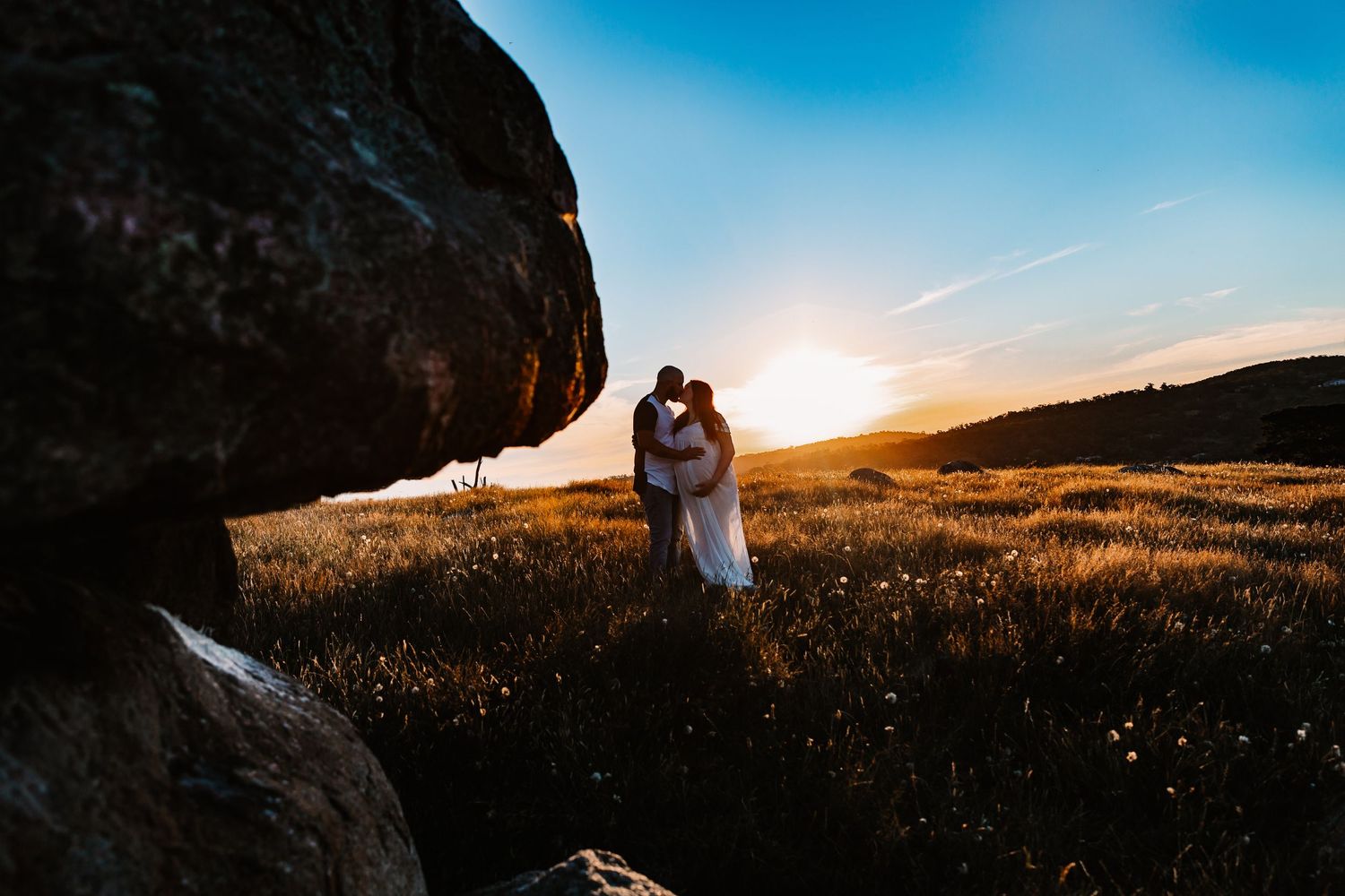 A couple in a field at sunset, kissing and embracing near some granite boulders