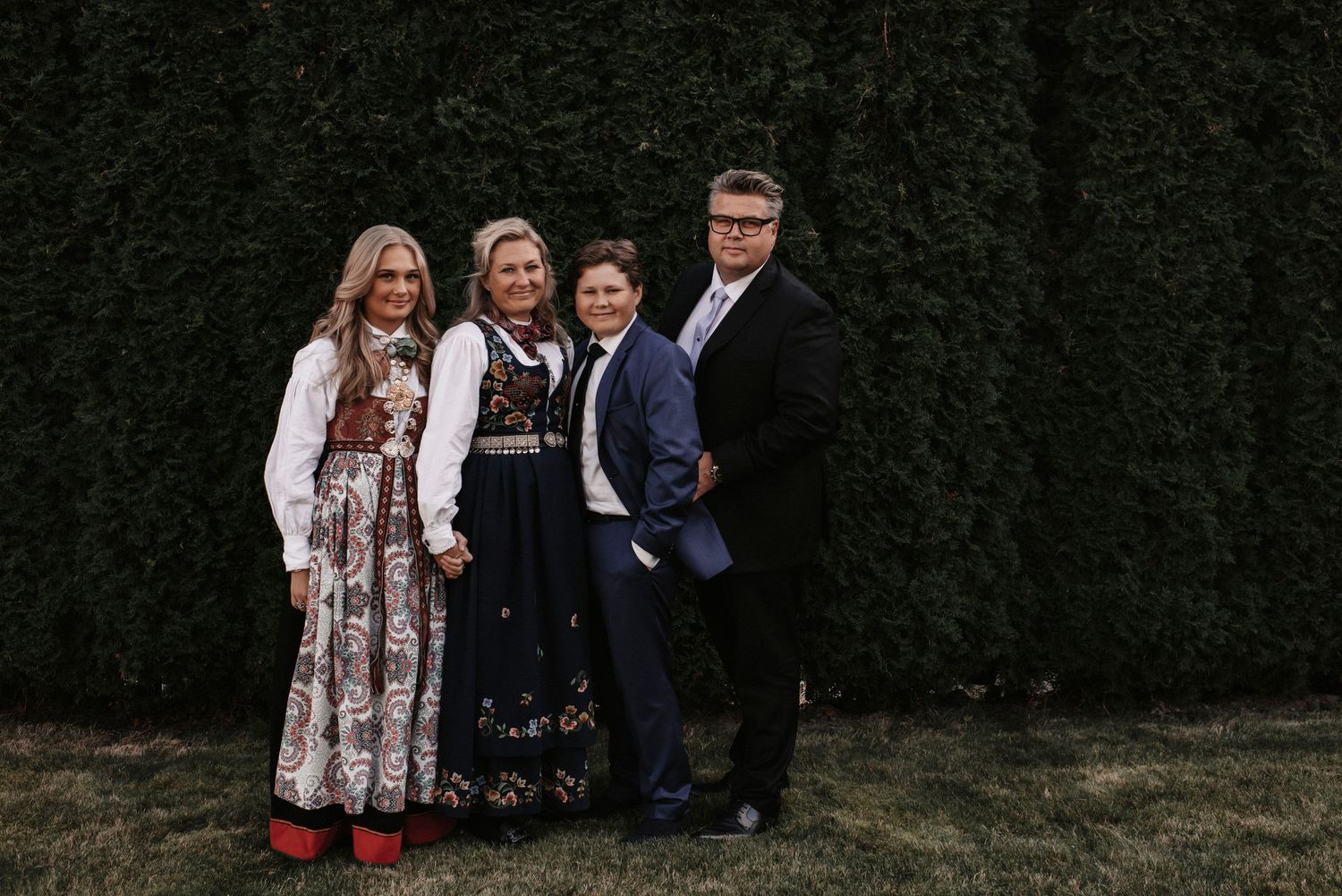 Family posing in mix of traditional Norwegian clothing and formal wear against dark green foliage.