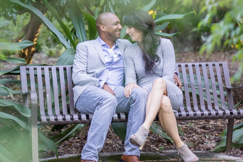 A couple sits together on a wooden bench surrounded by lush tropical plants in a garden setting.