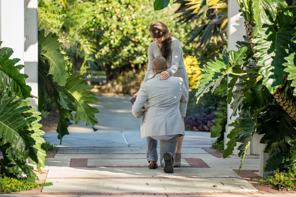 Romantic proposal scene on garden walkway surrounded by tropical plants.