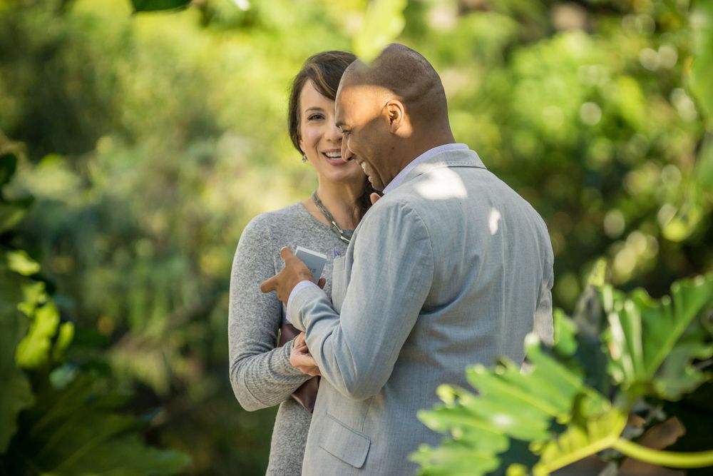 Two people share an intimate moment surrounded by tropical foliage in a garden setting.