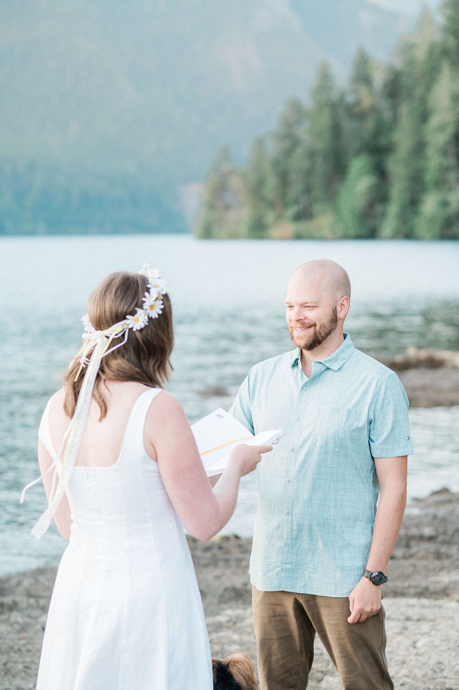A bride and groom share a romantic moment by a scenic mountain lake with evergreen trees in background.