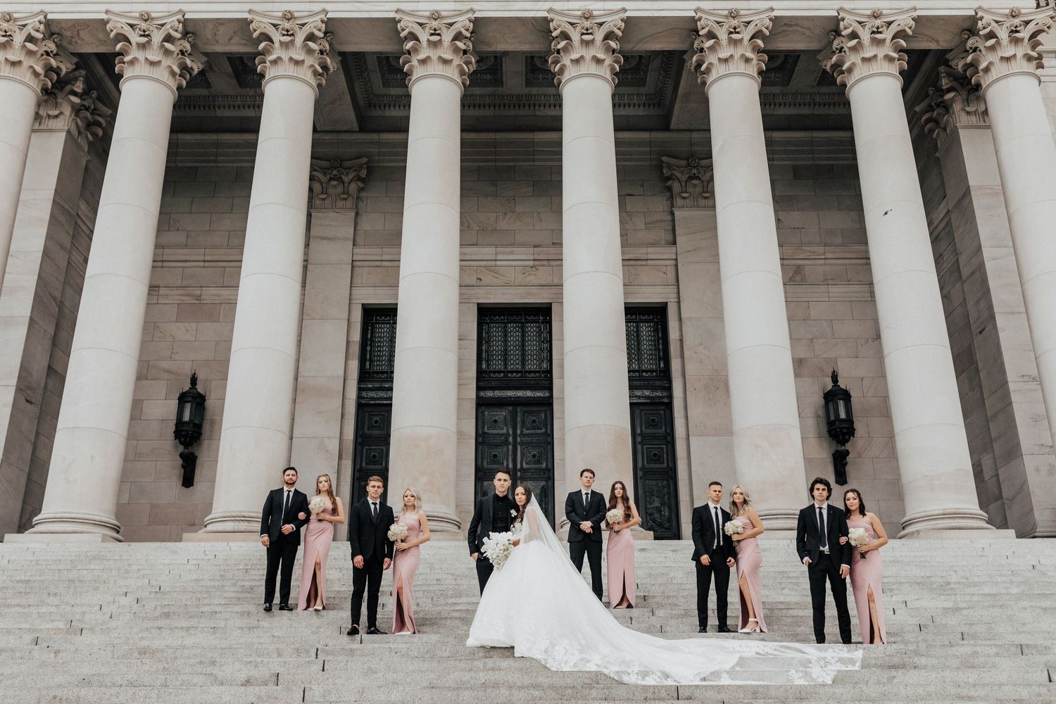 Wedding party poses elegantly on the steps of a grand neoclassical building with white stone columns.