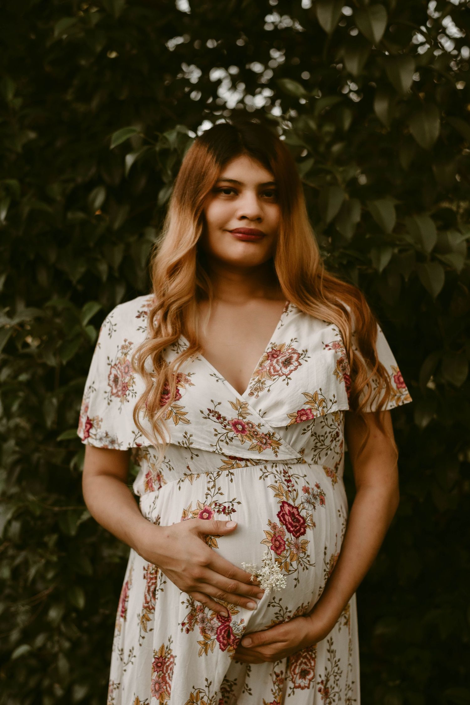 A maternity photo showing someone wearing a white floral dress against a background of green leaves.