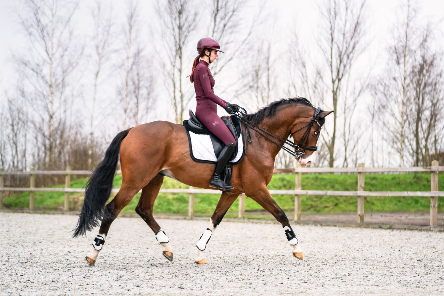 Elegant dressage rider in burgundy riding a bay horse through an outdoor arena.