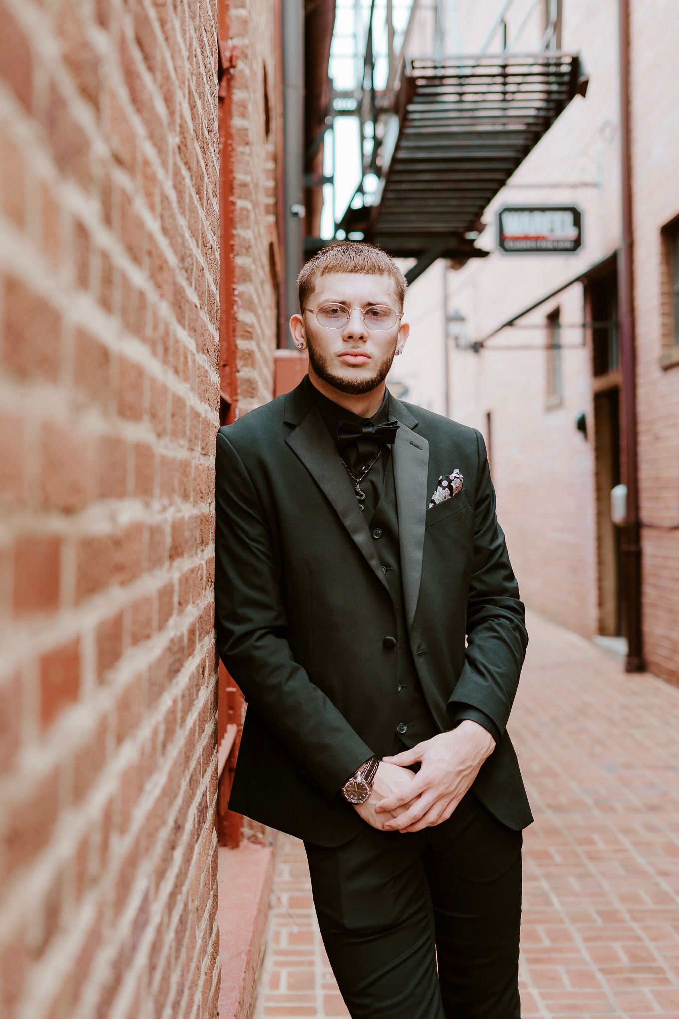 Person in black formal attire poses against brick walls in an urban alleyway setting.