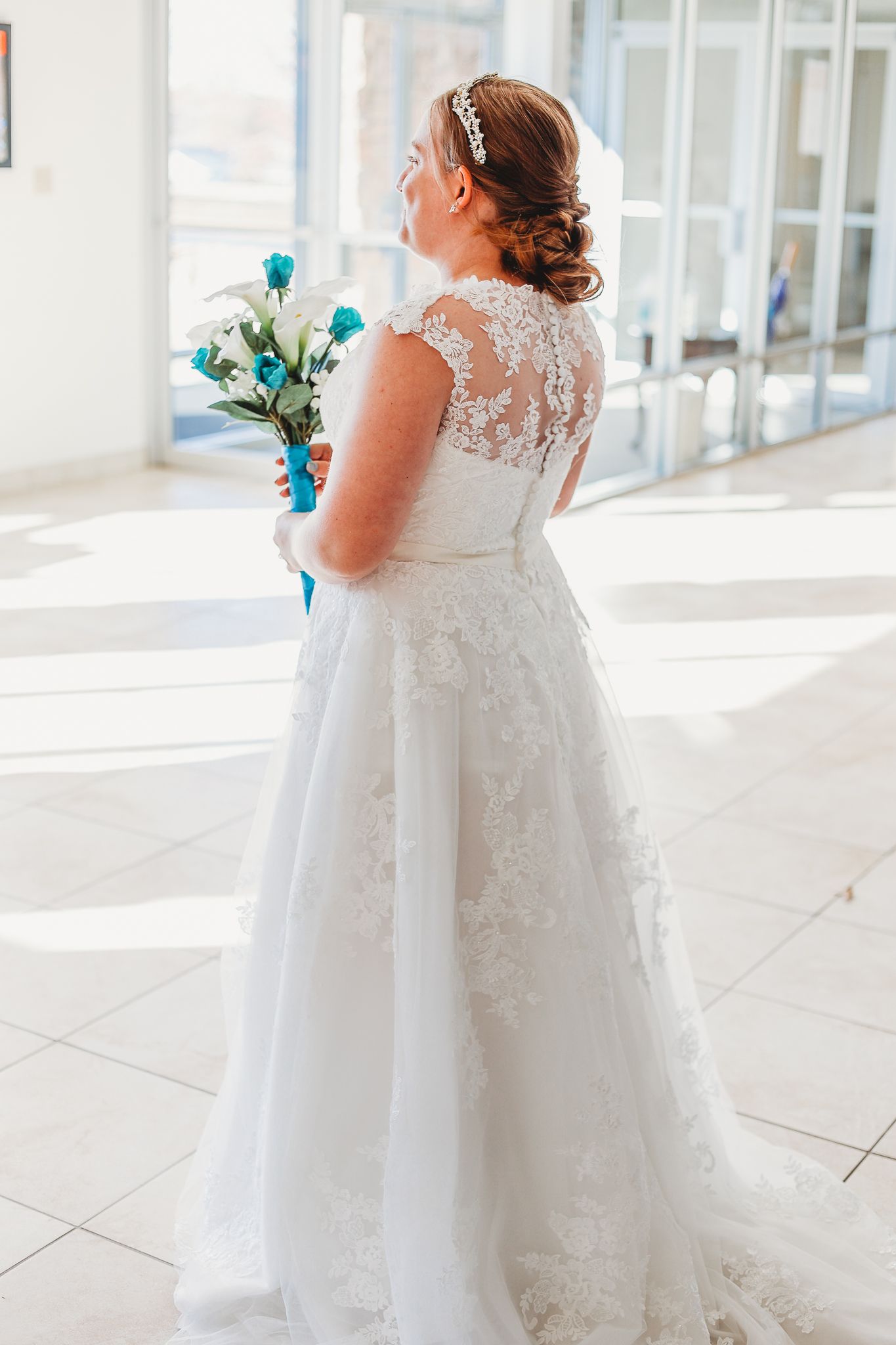 A bride in a white lace wedding dress with illusion back holds a turquoise bouquet while standing in a sunlit venue.