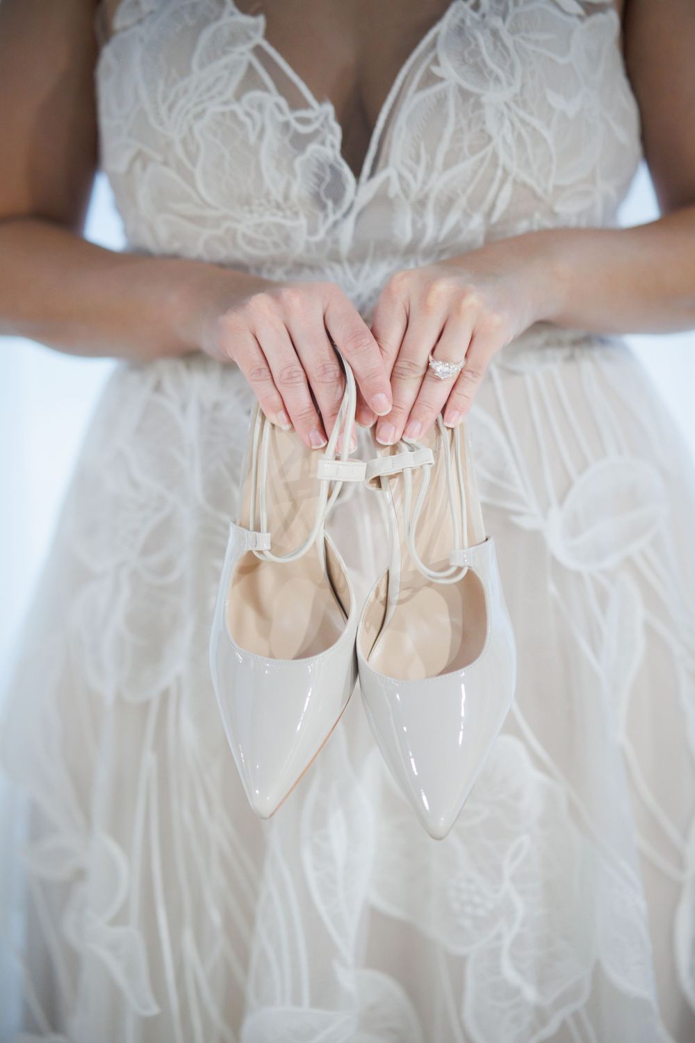 Bride holding white patent leather wedding shoes against her lace dress.