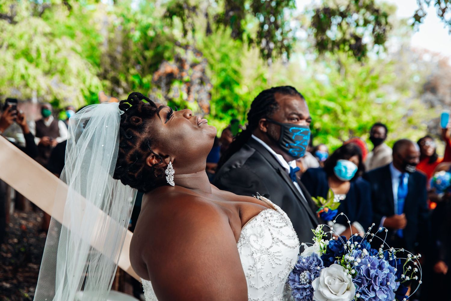 Emotional bride in lace gown and veil reacts during outdoor wedding ceremony with masked guests behind her.