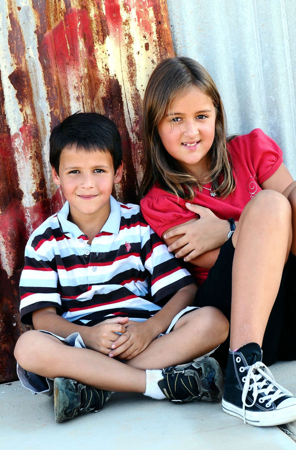Two children sit together against a rustic metal wall background.