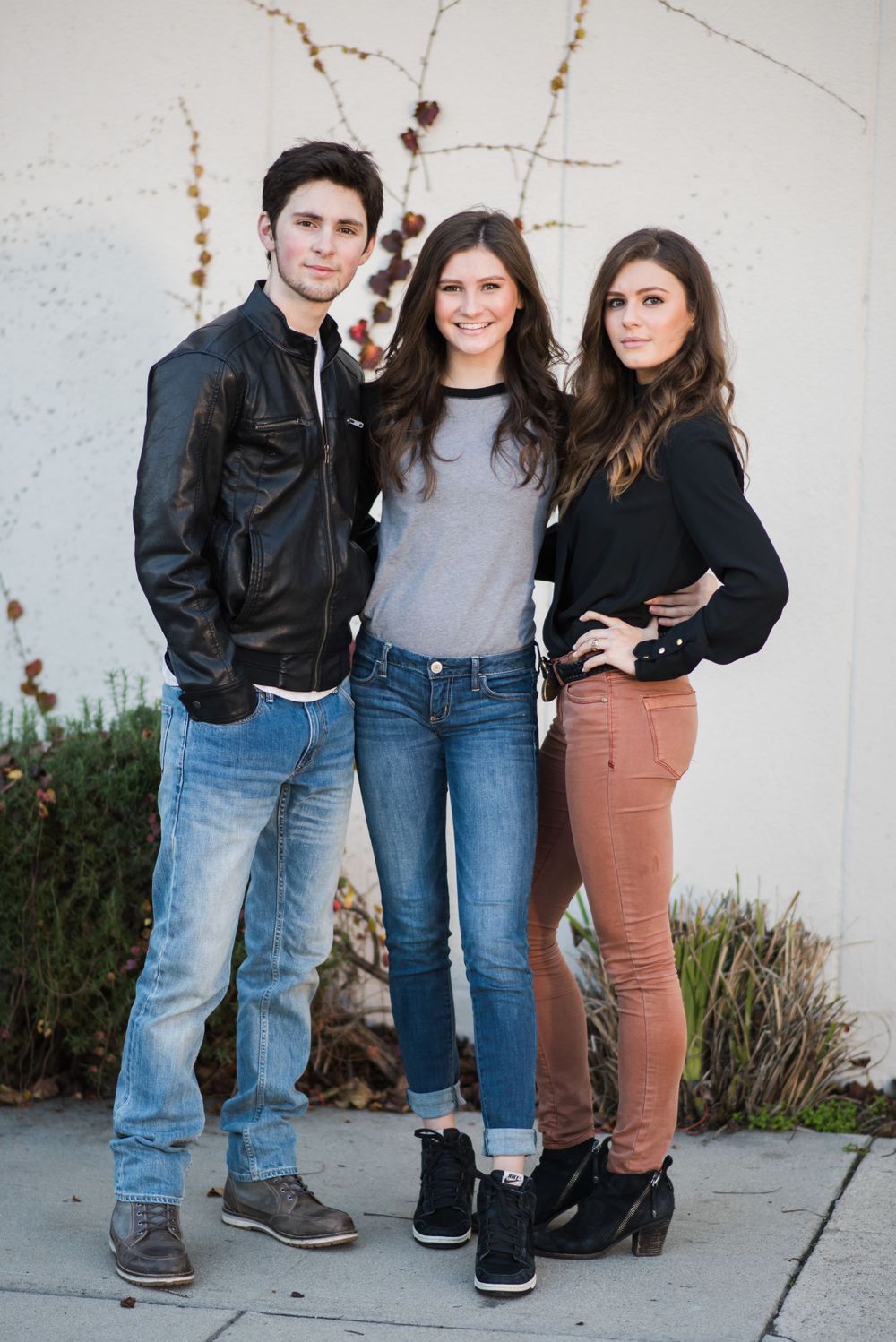 Three young people in casual wear posing against white wall with winter branches.