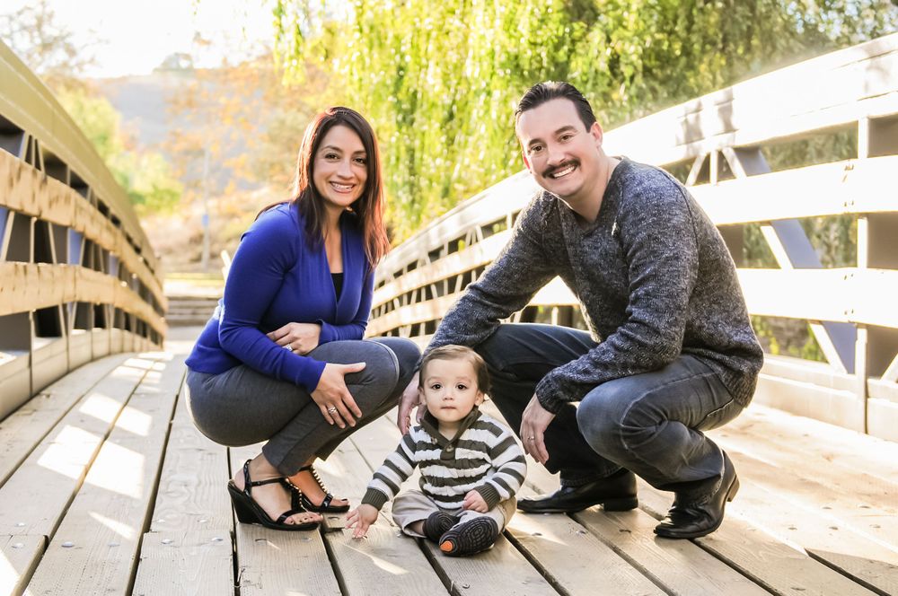 Family sitting together on wooden bridge with willow trees in background.
