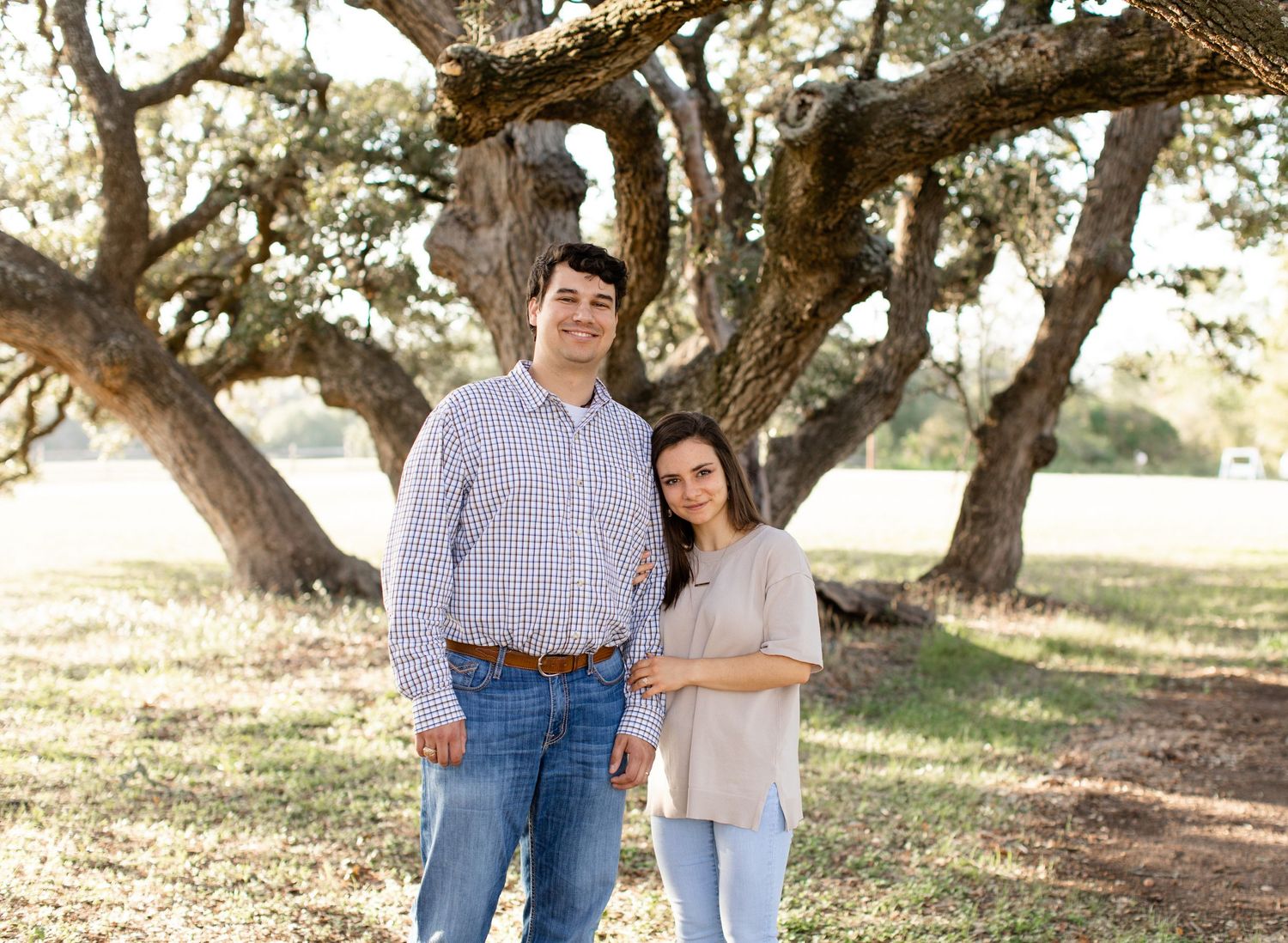 A couple stands together under sprawling oak trees in a grassy field.