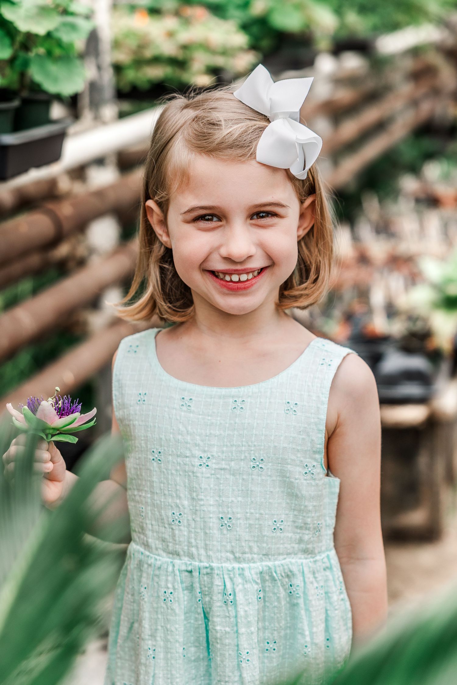 A figure in a mint green dress holds a purple flower in a greenhouse setting.