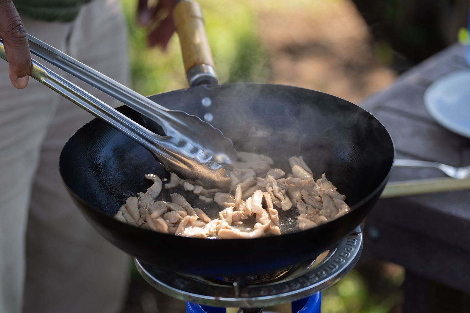 Chef cooking meat in cast iron pan over outdoor flame.