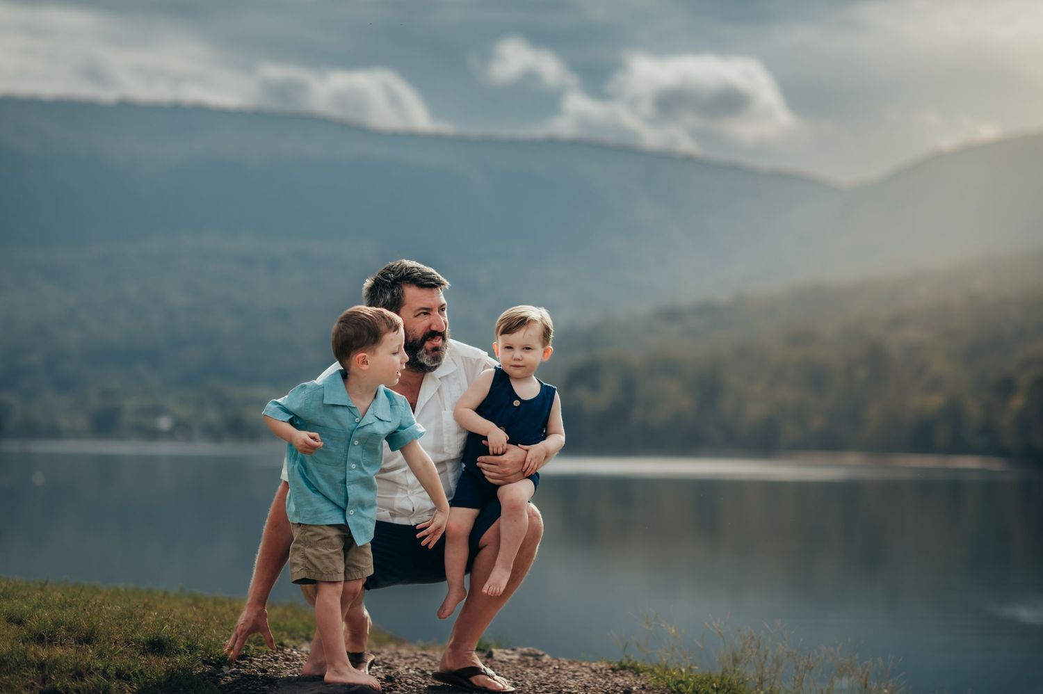 Dad crouches with his 2 young sons as the eldest throws rocks into the river at the base of Raccoon Mountain