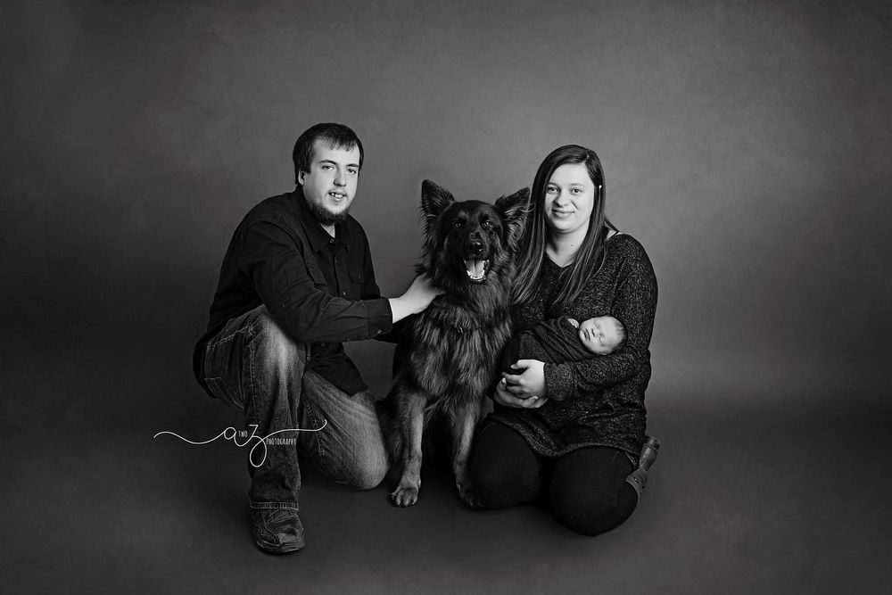 A black and white portrait of a couple with their German Shepherd dog in a studio setting.