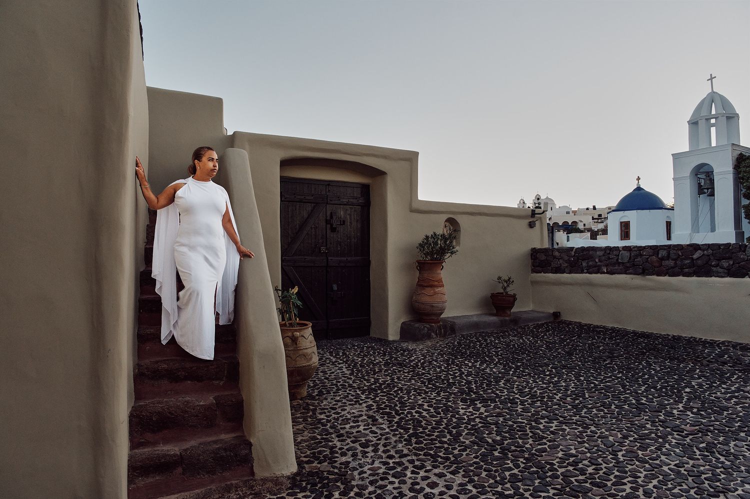Woman in white outfit on steps of Mediterranean building with blue-domed church in Santorini, Greece.