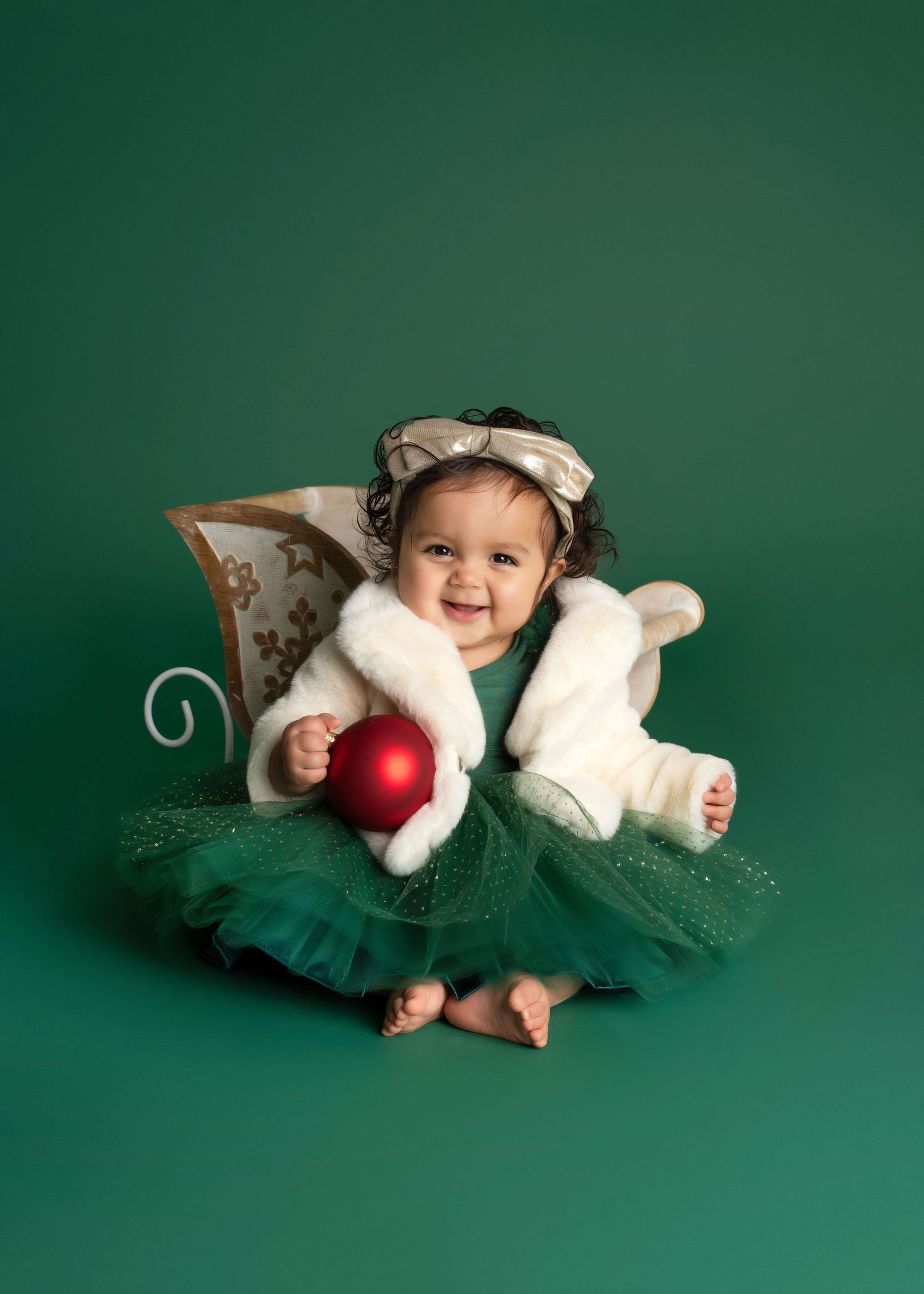 in winter onederland, a baby girl smiles while wearing a green dress, cream bow, and cream fur jacket