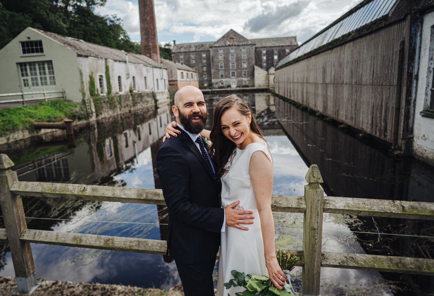 A wedding couple embraces on a wooden dock with industrial buildings in the background.