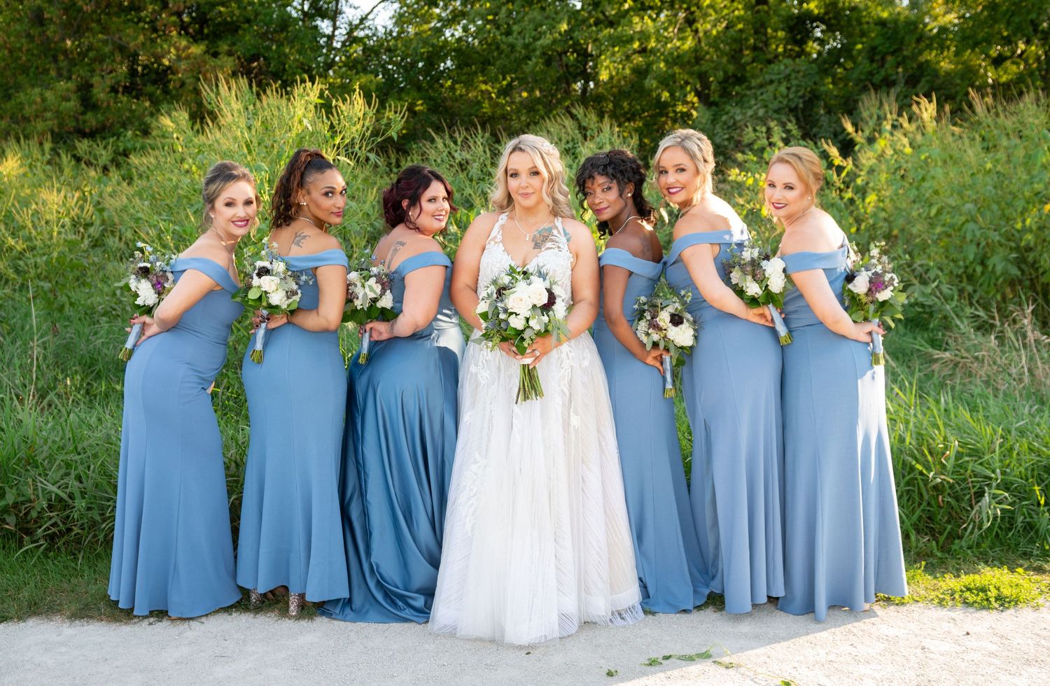 Bridesmaids in dusty blue dresses pose with bouquets outdoors in a garden setting.