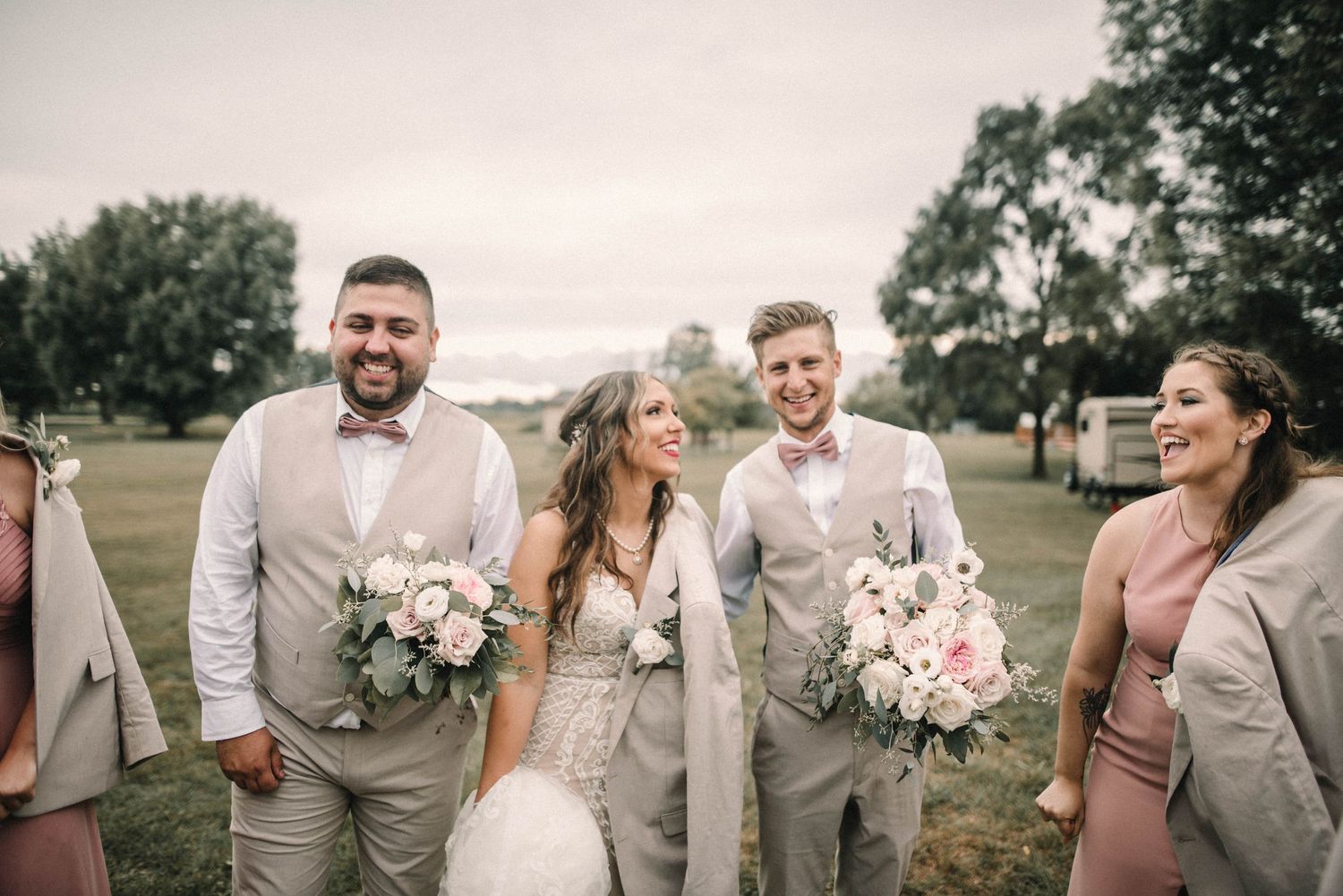 A wedding party in light gray attire poses together outdoors holding pink bouquets.