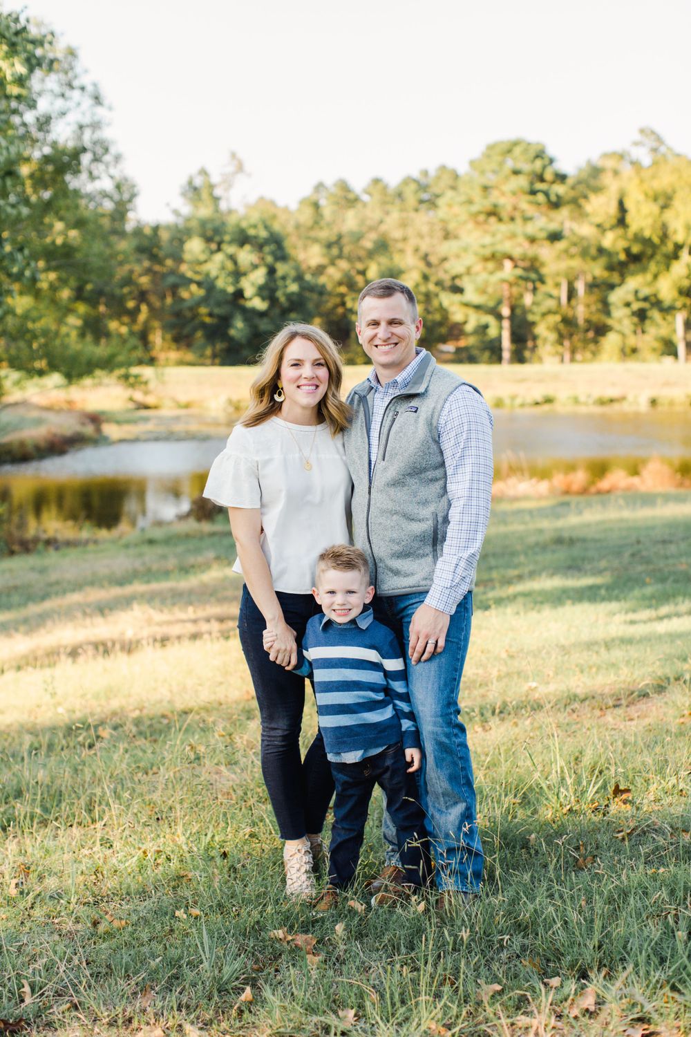 Family of three poses together on grassy hill with lake in background.