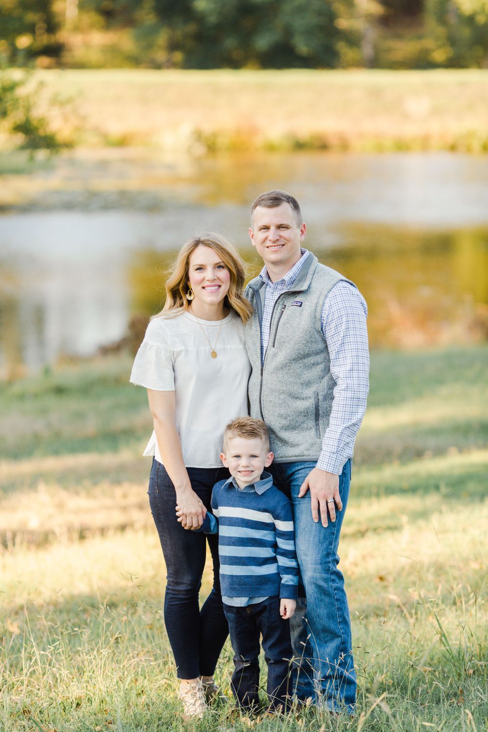 Casual family portrait outdoors with pond and autumn foliage in background.