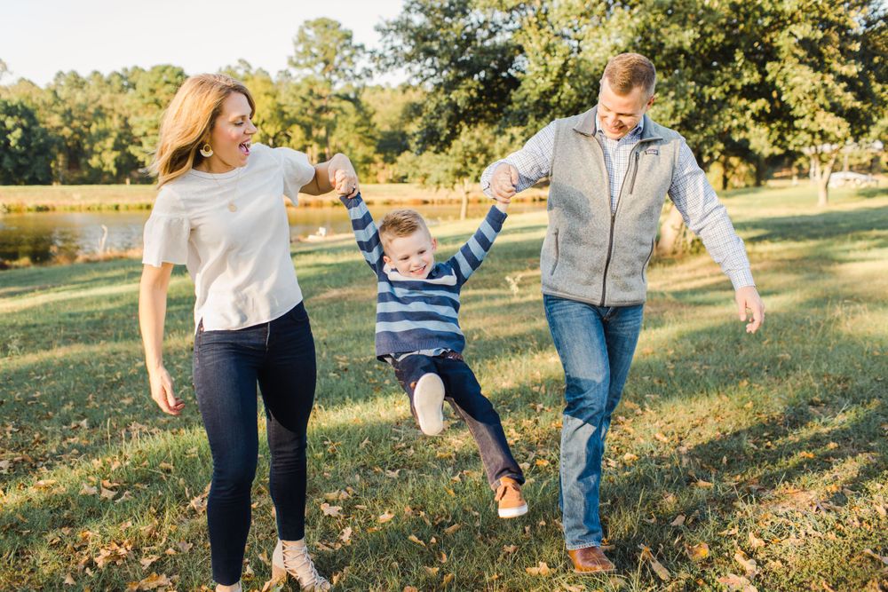 A family plays together in a sunlit park, swinging a young child between them on an autumn afternoon.