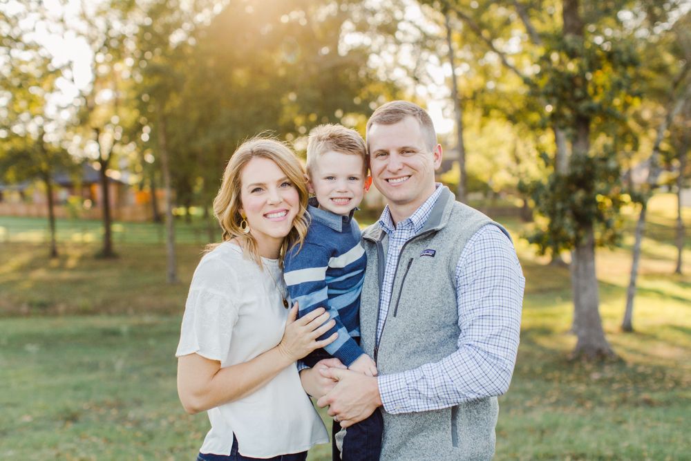 A family poses together outdoors in casual attire against a backdrop of autumn trees.