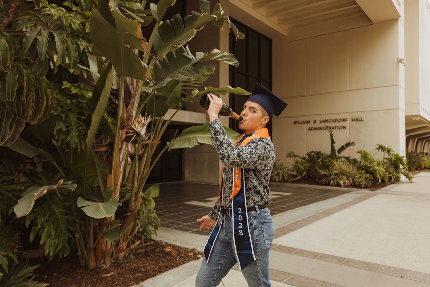 Graduate in blue cap and gown celebrating outdoors near tropical plants.