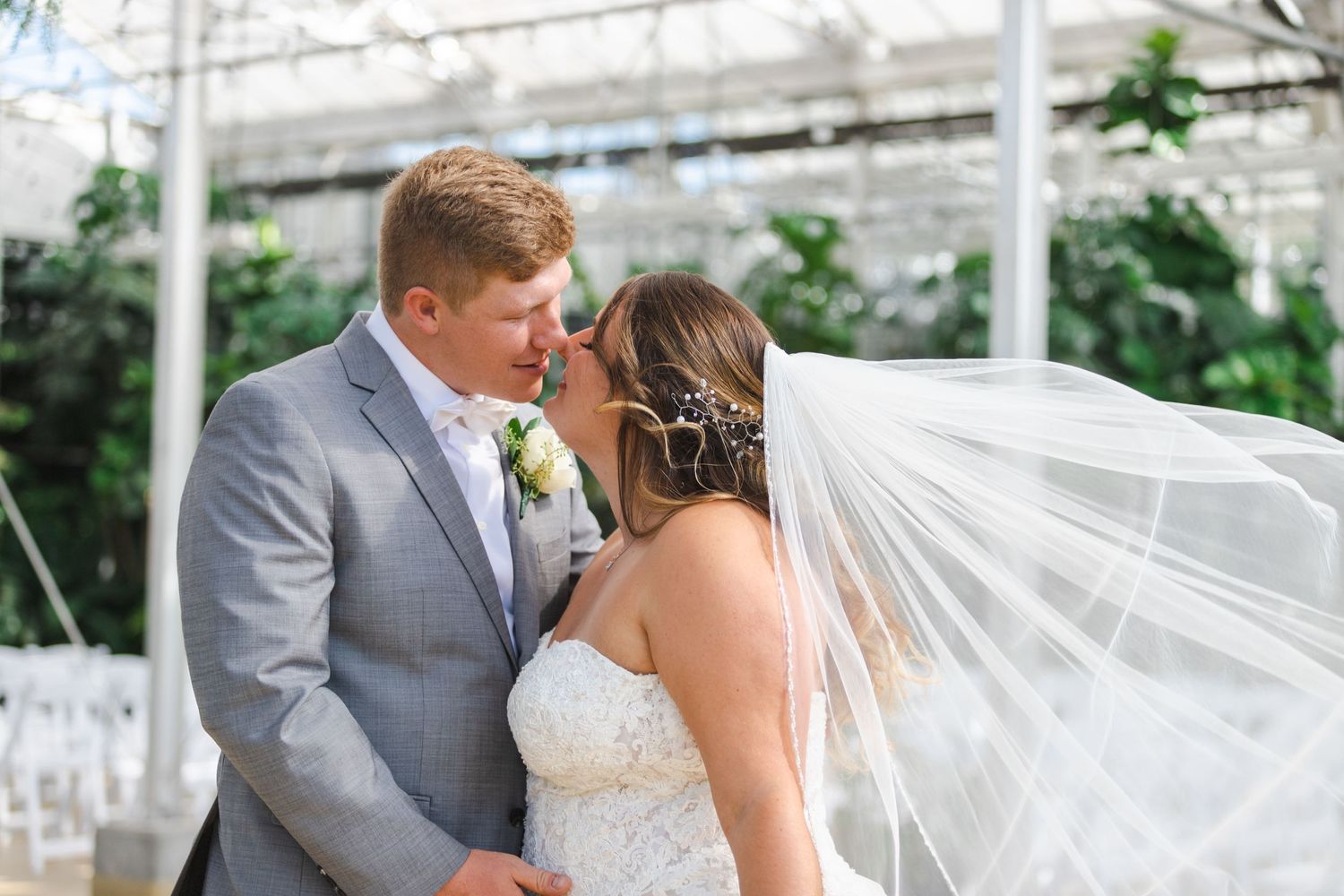 Newlyweds sharing a romantic kiss in a greenhouse with a flowing white veil.