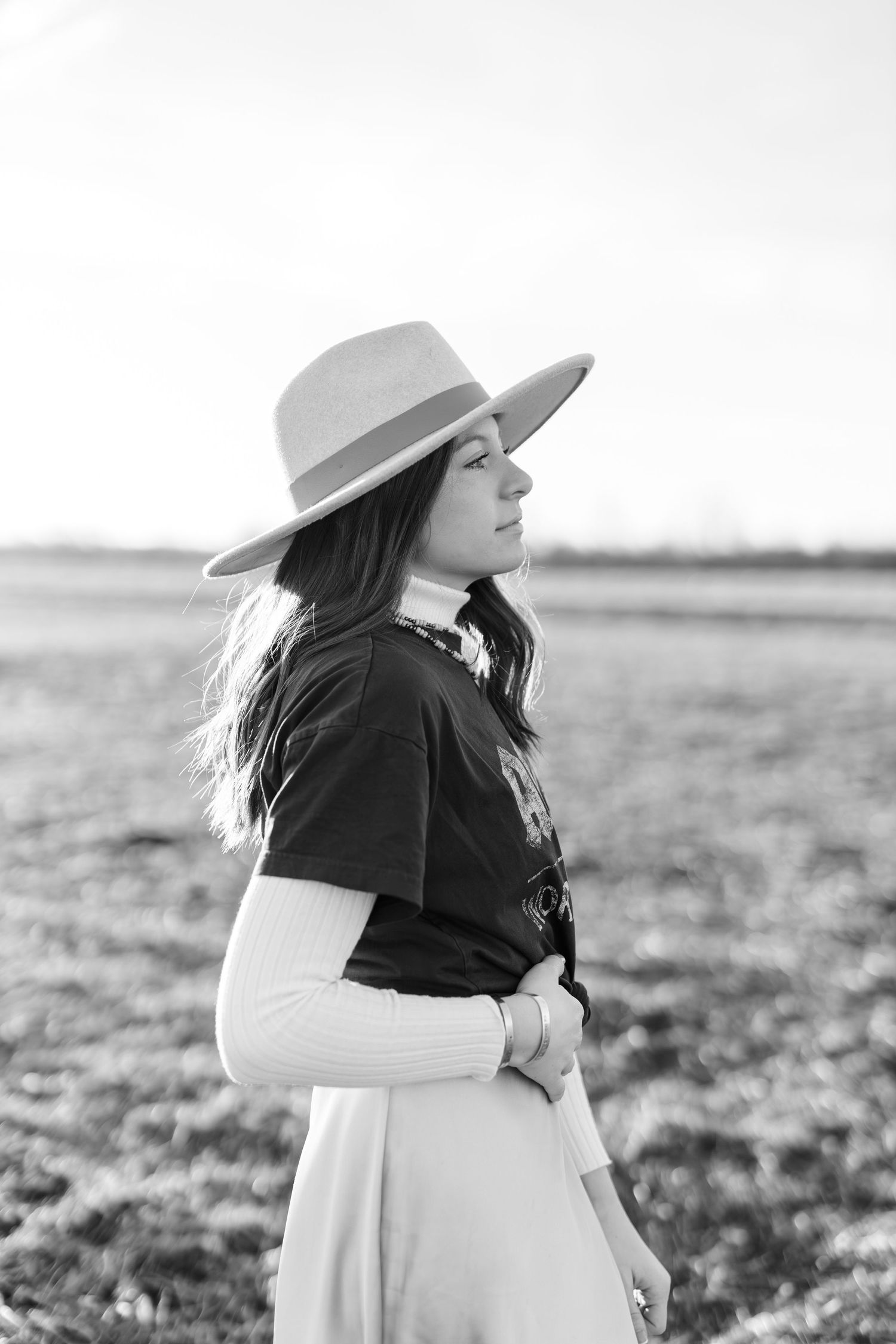Black and white photo of a young woman in a wide-brim hat and dark shirt standing in an open field.