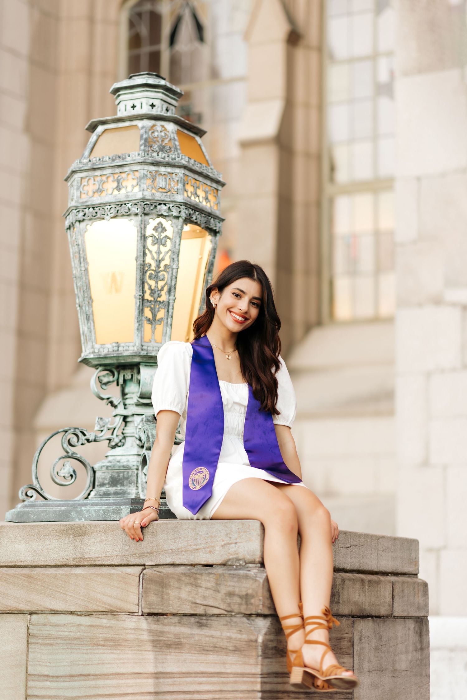 Graduate in white dress with purple sash sits by decorative lamp post.
