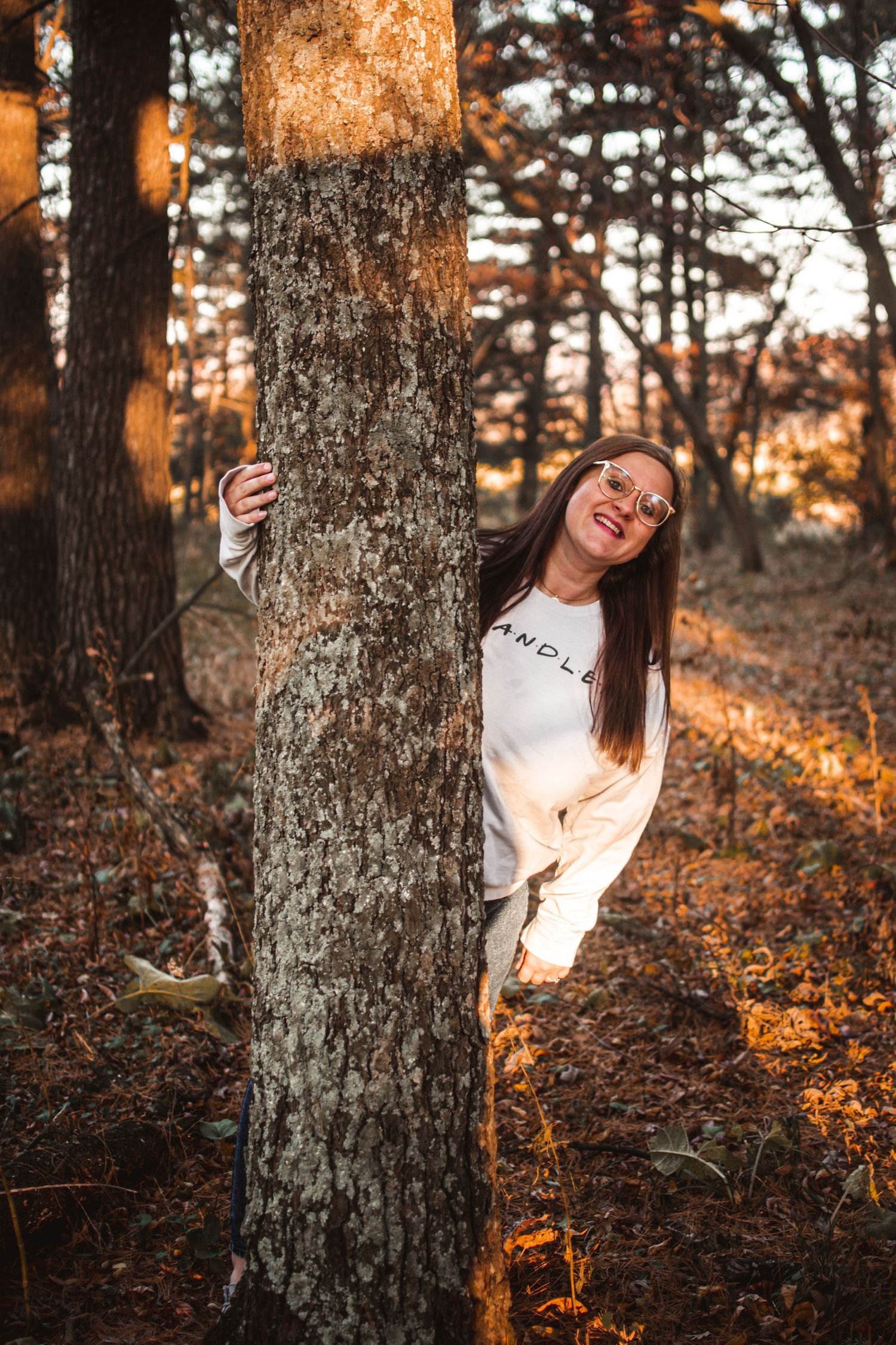 Person playfully peeking from behind a large tree trunk in an autumn forest.