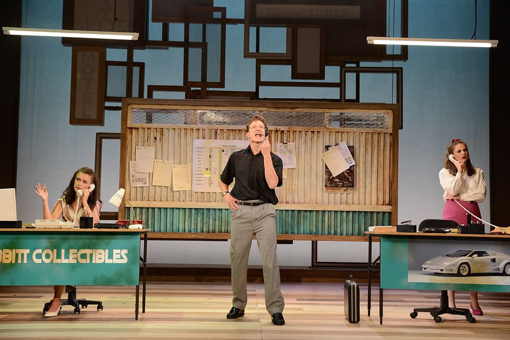 Three actors perform a scene in an office setting with retro furniture and decor.
